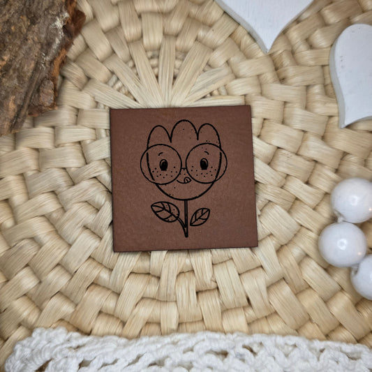 Kunstlederlabel „Buntes Glück“ mit verspielter Blumenillustration mit runder Brille, schwarz graviert auf braunem Kunstleder, ca. 4×4 cm