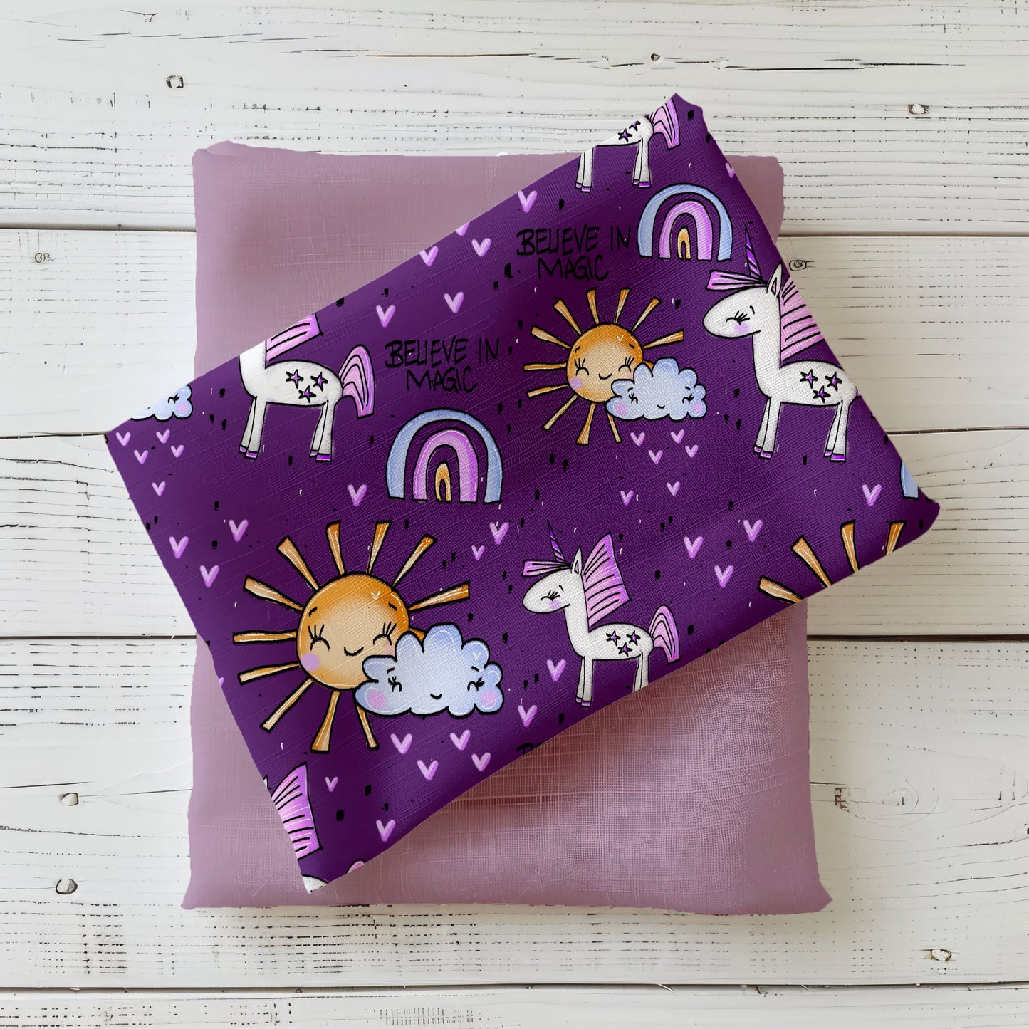 Mockup: Stoff „Believe in Magic – Einhorn“ in Lila mit weißen Einhörnern (rosa/lila Mähne), Regenbögen, Sonne und Wolke, vielen kleinen Herzchen und dem Schriftzug „Believe in Magic“; Stoffstück liegt diagonal auf fliederfarbenem Kombistoff, fotografiert auf weißer Holzfläche.