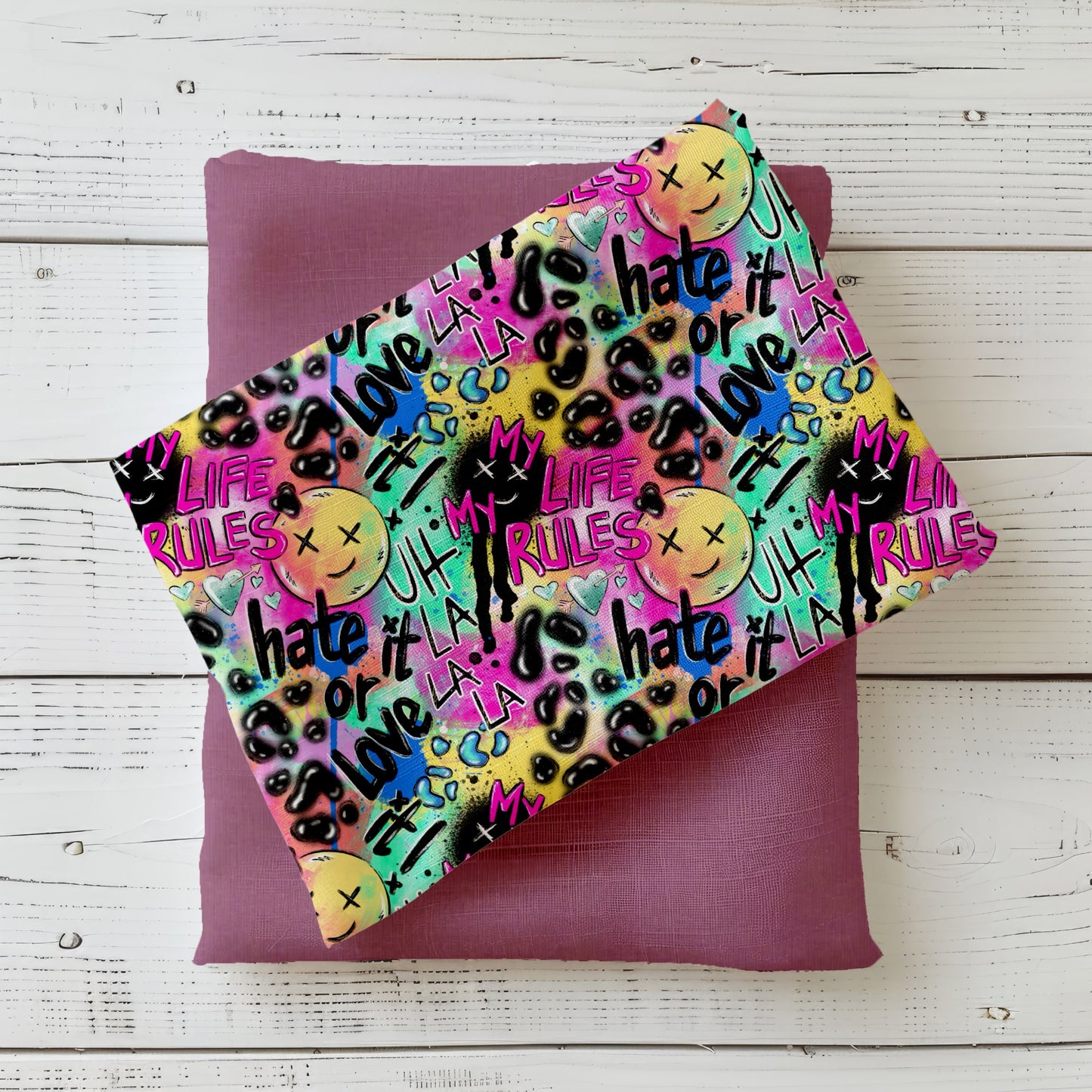Mockup: Stoff „Hate it or Love it“ – Graffiti-Schriftzüge, Smileys, Drips & Leo-Sprenkel in Neon-Pink, Lila, Türkis, Gelb & Lime auf dunklen Akzenten; Stoffkachel diagonal auf passendem Uni in Mauve/Lila als Kombi.