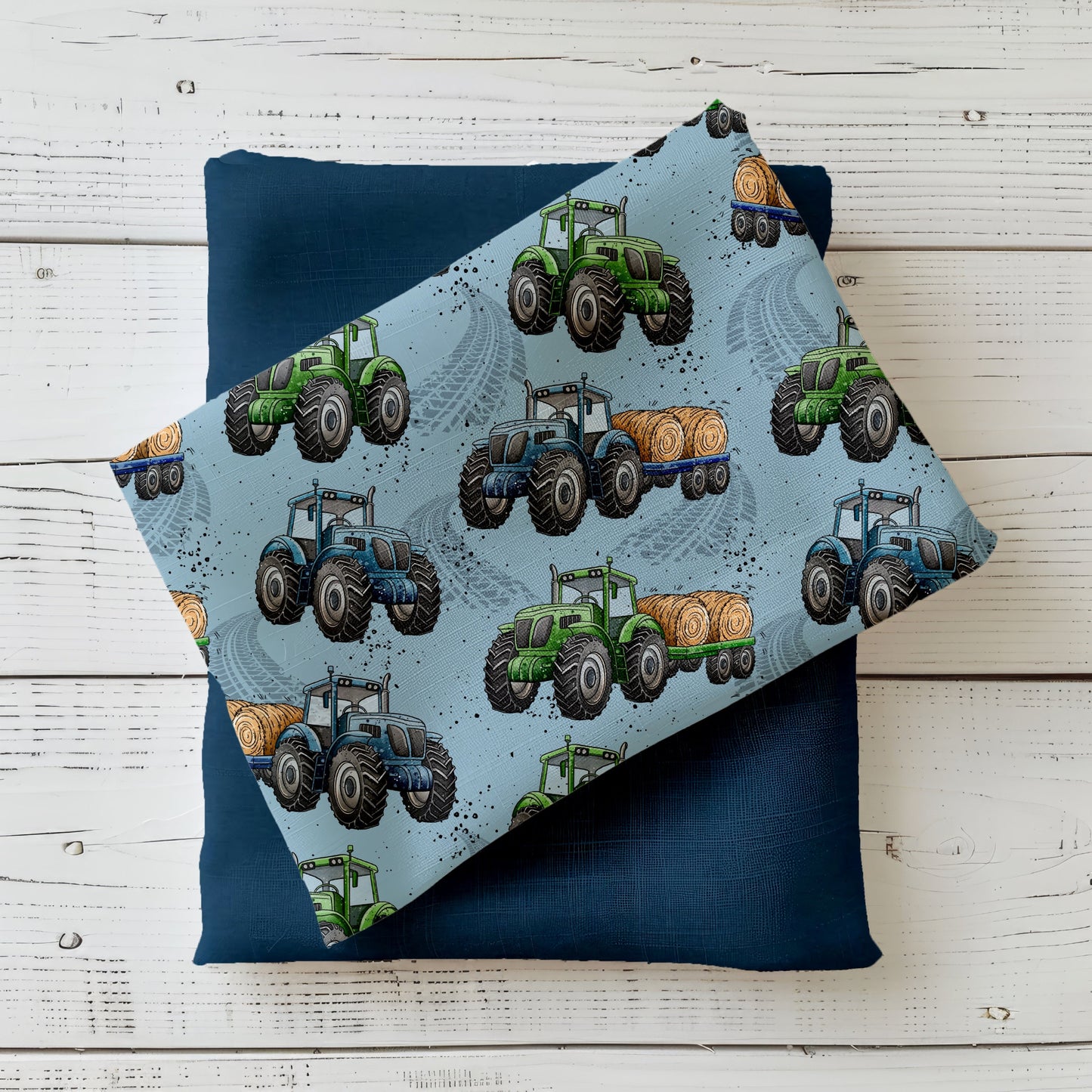 Mockup: Stoff „Trecker-Crew“ mit grünen und blauen Traktoren sowie Strohballen-Anhänger auf hellblauem Hintergrund mit Reifenspuren; aufgelegt auf dunkelblauem Kombistoff, fotografiert auf hellem Holz.