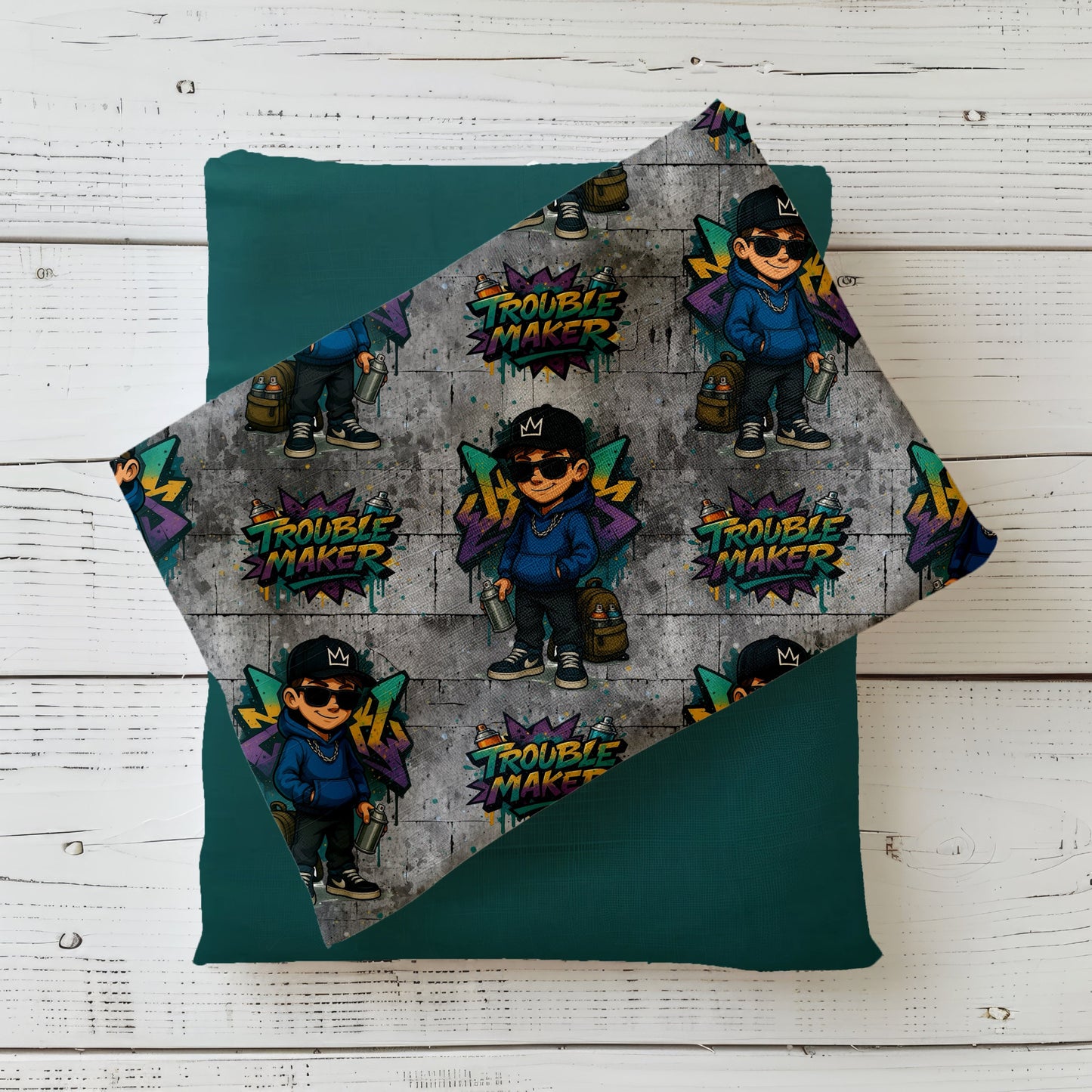Mockup eines gefalteten Stoffes mit dem „Trouble Maker“-Design: Wiederholendes Graffiti-Muster mit coolem Jungen und buntem Schriftzug auf grauer Betonwand – ideal für modische Kinderkleidung im Streetwear-Stil.