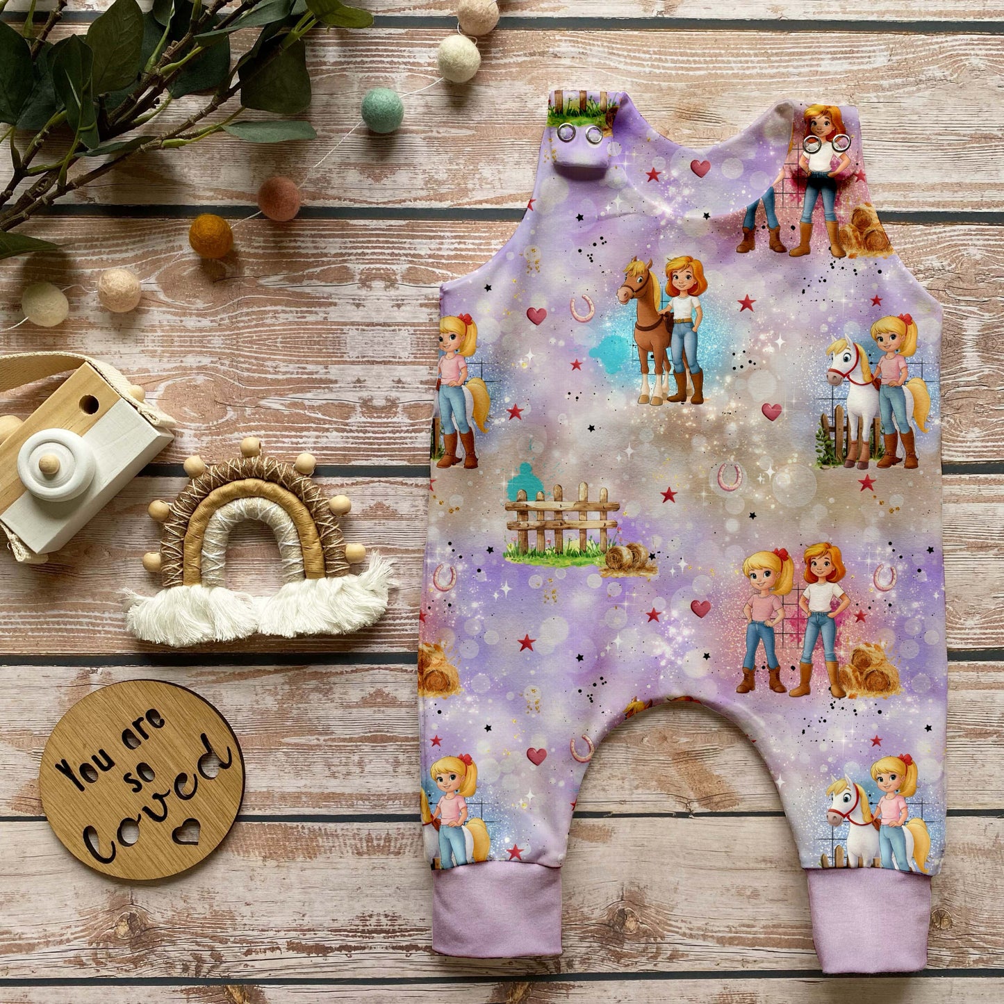 Handgemachter Baby-Strampler im Design „Zauberhafte Reiterinnen“ mit Reiterinnen, Pferden und Naturmotiven auf zartem Hintergrund sowie lavendelfarbenen Bündchen, arrangiert auf hellem Holz mit natürlichen Accessoires.