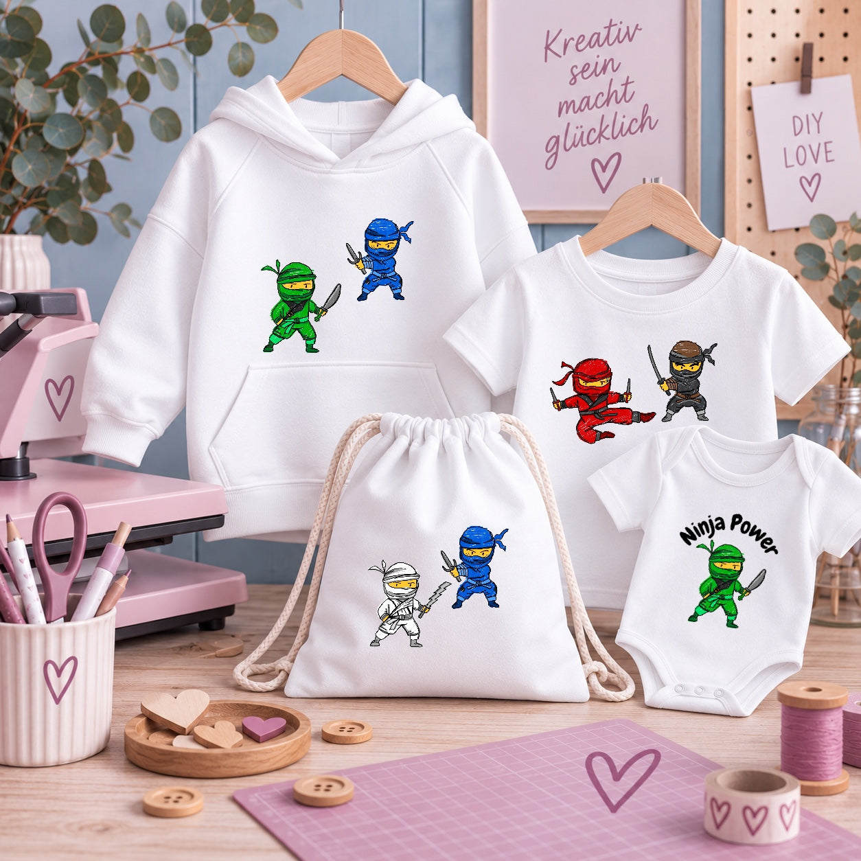 Weiße Kinderkleidung mit Ninja-Motive: Hoodie, T-Shirt, Beutel und Baby-Body. Dazwischen Bastelzubehör wie Schere, Knöpfe und Transferpresse vor hellblauer Wand mit Spruch „Kreativ sein macht glücklich“