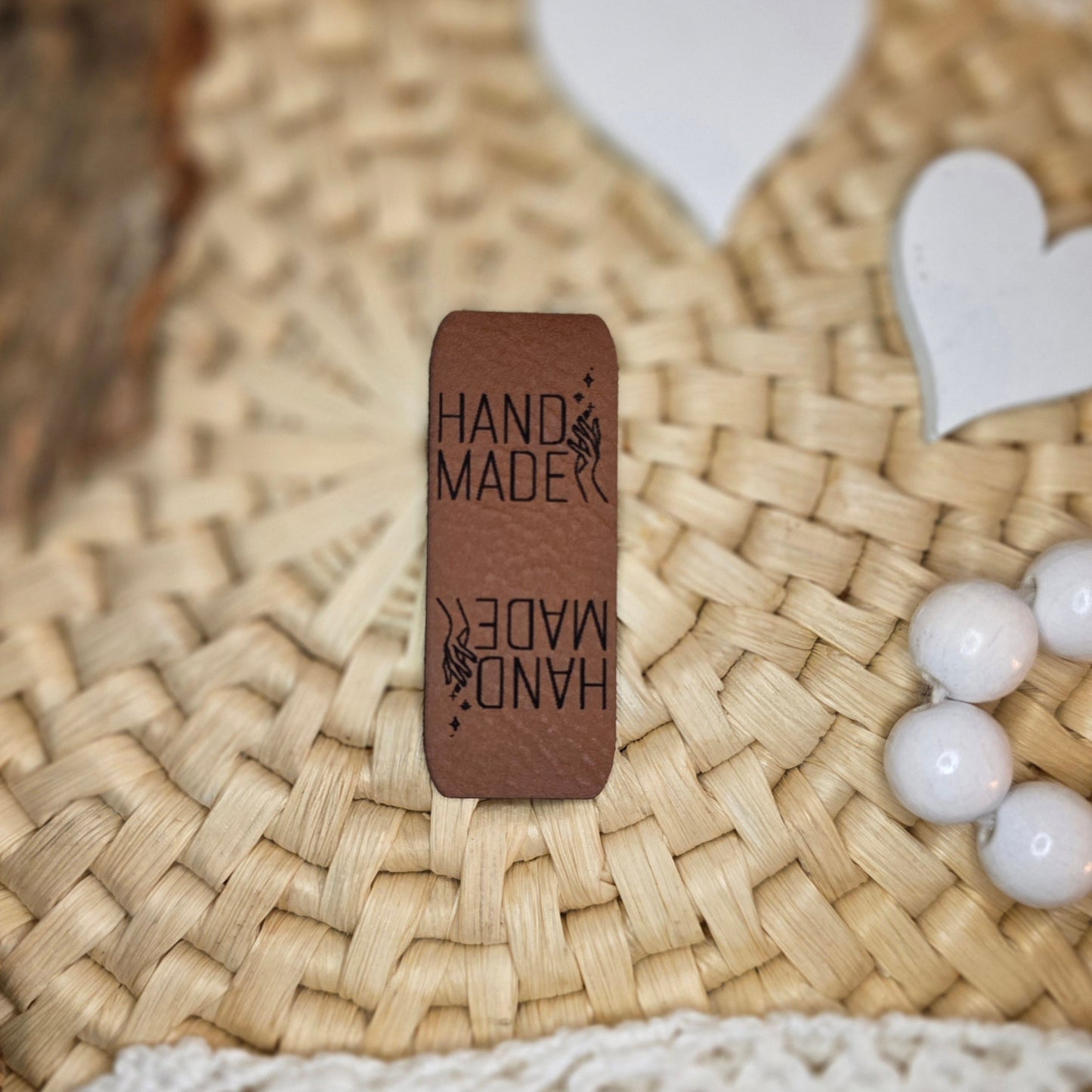 Knicklabel „Handmade“ aus braunem Kunstleder (5 × 2 cm), beidseitig graviert mit schwarzer Schrift und Handmotiv, dekorativ platziert auf Bastuntergrund mit Holzrinde, weißen Dekoherzen und Häkelspitze – elegantes Label für handgemachte Kleidung, Accessoires und Unikate.