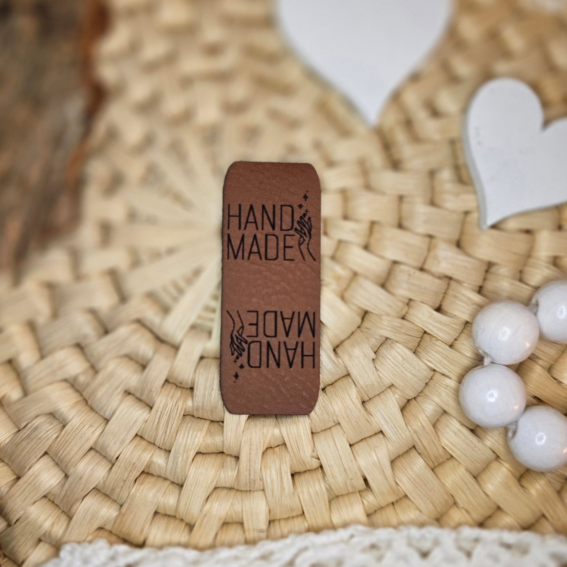 Knicklabel „Handmade“ aus braunem Kunstleder (5 × 2 cm), beidseitig graviert mit schwarzer Schrift und Handmotiv, dekorativ platziert auf Bastuntergrund mit Holzrinde, weißen Dekoherzen und Häkelspitze – elegantes Label für handgemachte Kleidung, Accessoires und Unikate.