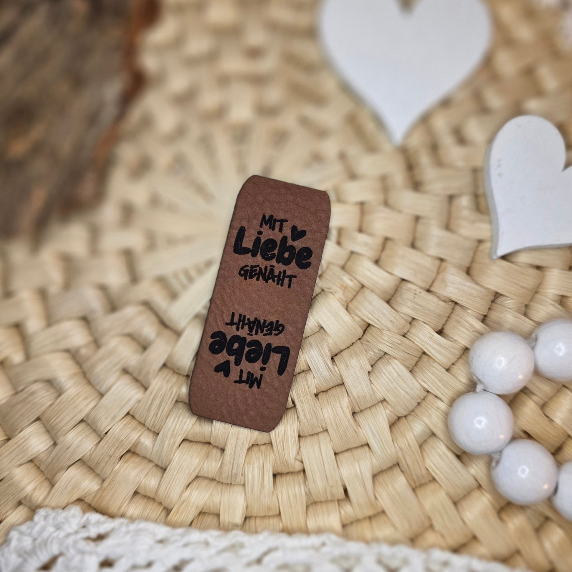 Knicklabel „Mit Liebe genäht“ aus braunem Kunstleder (5 × 2 cm), beidseitig graviert mit schwarzer Schrift und kleinen Herzsymbolen, dekorativ auf Bastuntergrund mit Holzrinde, weißen Dekoherzen und Häkelspitze arrangiert – liebevolles Einnählabel für selbstgenähte Kleidung und Accessoires.
