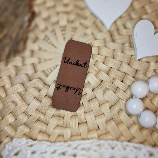Knicklabel „Unikat“ aus braunem Kunstleder (5 × 2 cm), beidseitig graviert mit schwarzer Schrift, dekorativ auf Bastuntergrund mit Holzrinde, weißen Dekoherzen und Häkelspitze arrangiert – stilvolles Label für selbstgenähte Einzelstücke und handgemachte Unikate.