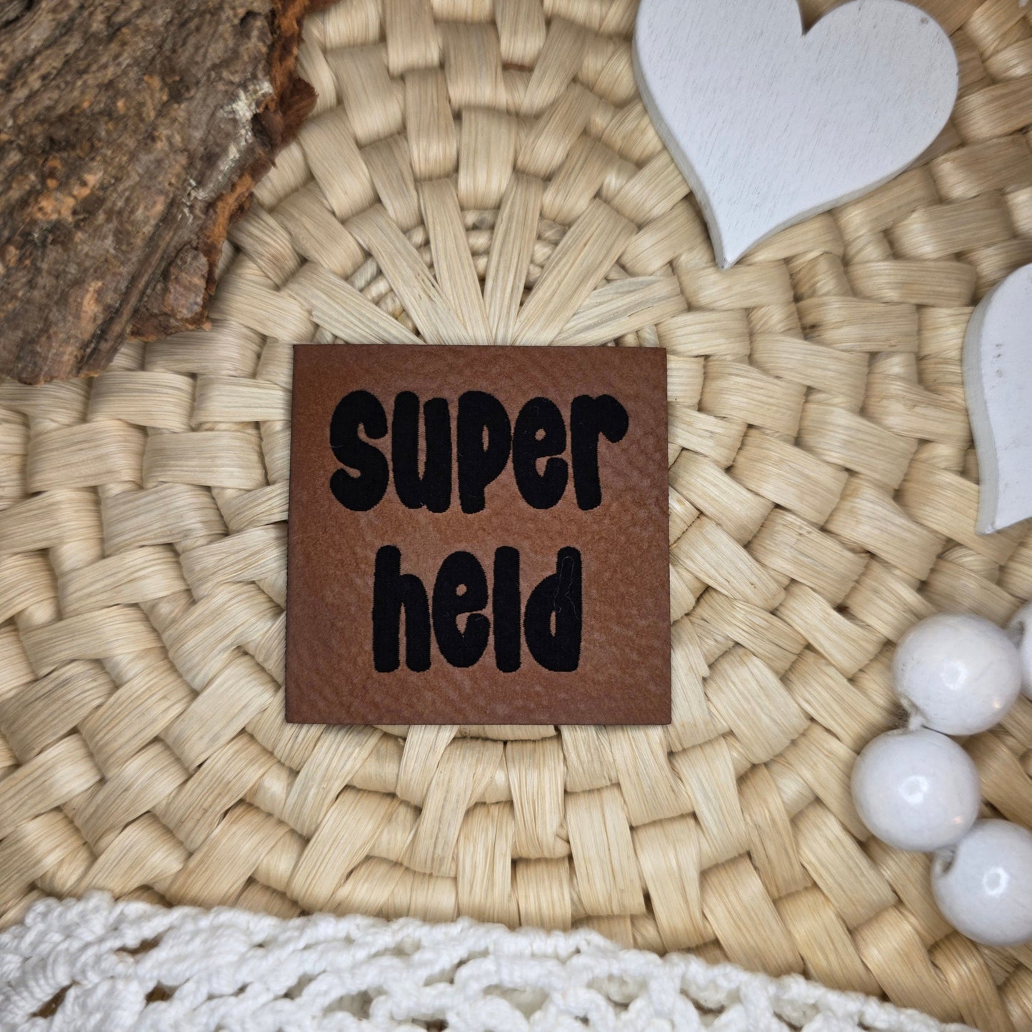 Quadratisches Kunstlederlabel „Superheld“ aus braunem Kunstleder (4 × 4 cm) mit schwarzer Gravur, dekorativ arrangiert auf Bastuntergrund mit Holzrinde, weißen Dekoherzen und Häkelspitze – ein kraftvolles Statement-Label für selbstgenähte Kinderkleidung und Accessoires.