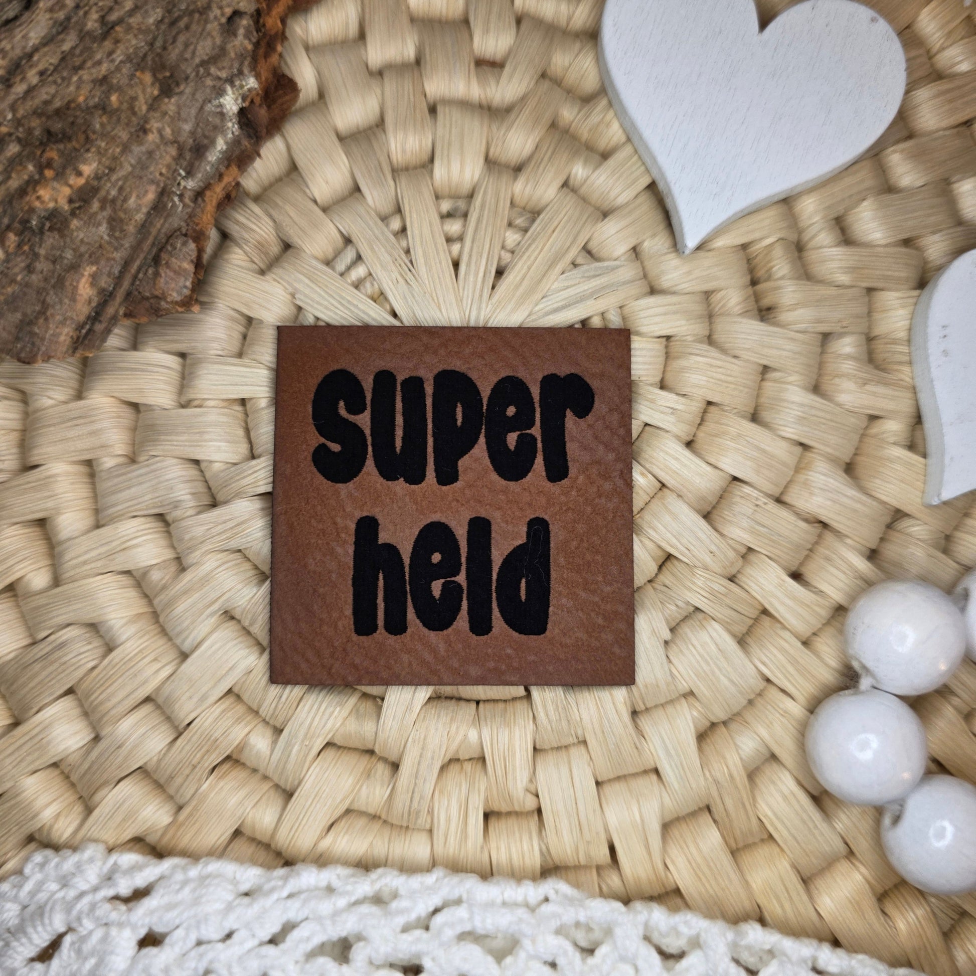 Quadratisches Kunstlederlabel „Superheld“ aus braunem Kunstleder (4 × 4 cm) mit schwarzer Gravur, dekorativ arrangiert auf Bastuntergrund mit Holzrinde, weißen Dekoherzen und Häkelspitze – ein kraftvolles Statement-Label für selbstgenähte Kinderkleidung und Accessoires.