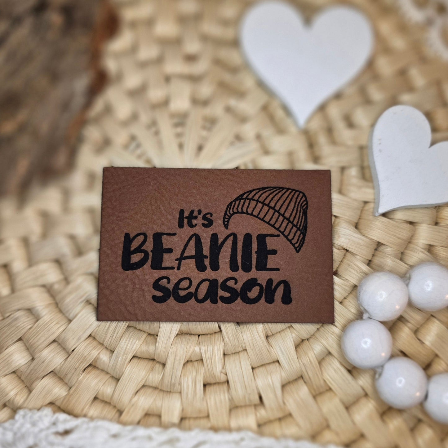 Kunstlederlabel „It’s Beanie Season“ aus braunem Kunstleder (6 × 4 cm) mit schwarzer Gravur und Mützen-Illustration, dekorativ auf Bastuntergrund mit Holzrinde, weißen Dekoherzen und Häkelspitze arrangiert – stilvolles Label für selbstgenähte Beanies, Stirnbänder und Winterkleidung.