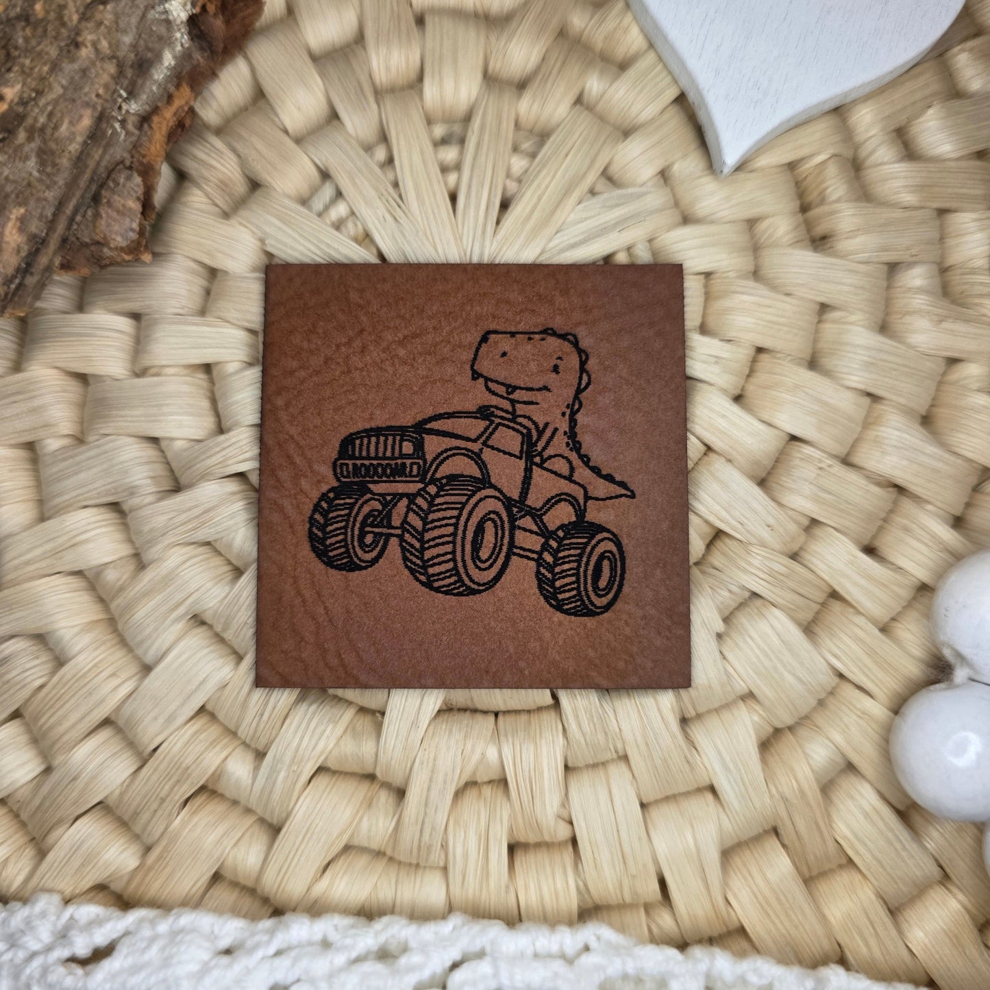 Quadratisches Kunstlederlabel „Dino Truck“ aus braunem Kunstleder (4 × 4 cm) mit schwarzer Gravur – handgezeichnetes Katzengold-Design mit Dino im Monstertruck, dekorativ auf Bastuntergrund mit Holzrinde, weißen Dekoherzen und Häkelspitze arrangiert – perfekt als Detail für selbstgenähte Kinderkleidung und Accessoires.