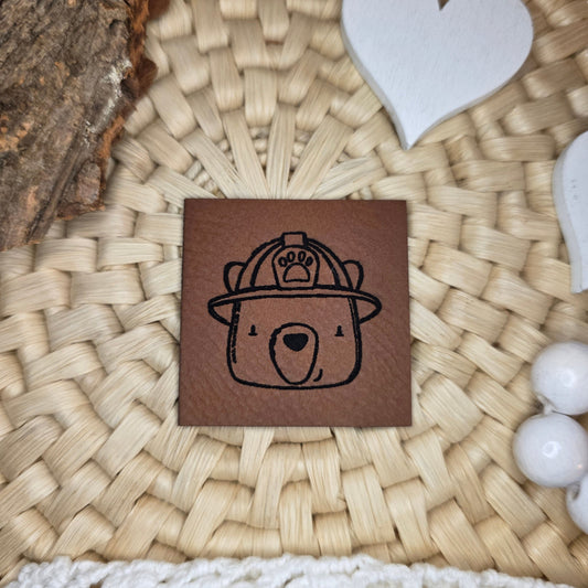 Quadratisches Kunstlederlabel „Fire Crew“ aus braunem Kunstleder (4 × 4 cm) mit schwarzer Gravur – zeigt den Bären mit Feuerwehrhelm aus der Fire-Crew-Kollektion von Katzengold-Designs; arrangiert auf Bastuntergrund mit Holzrinde, weißen Dekoherzen und Häkelspitze – perfektes Label für selbstgenähte Kinderkleidung im Feuerwehr-Look.