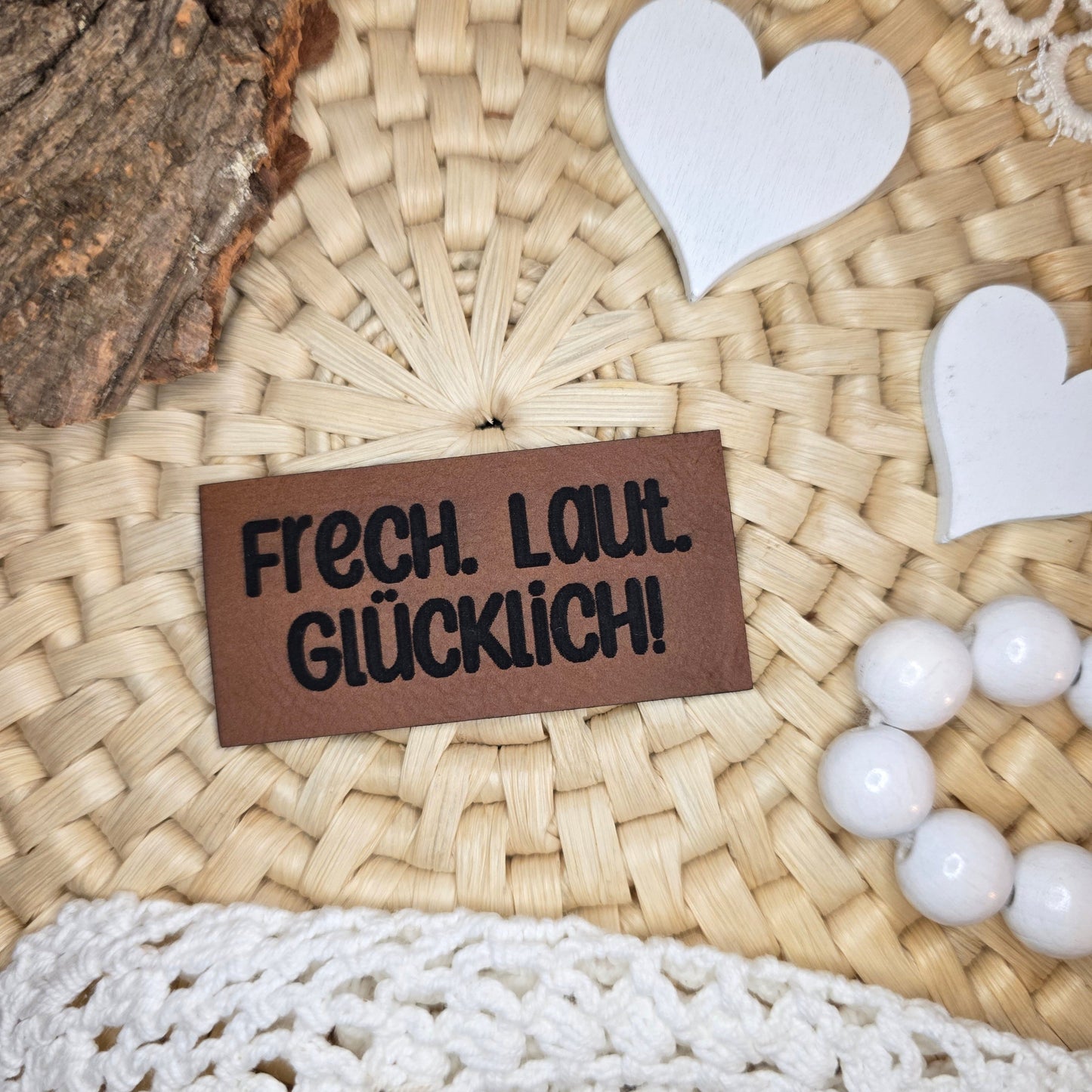 Kunstlederlabel „Frech. Laut. Glücklich!“ aus braunem Kunstleder (6 × 3 cm) mit schwarzer Gravur, dekorativ arrangiert auf Bastuntergrund mit Holzrinde, weißen Dekoherzen und Häkelspitze – fröhliches Statement-Label für Kleidung, Taschen und Accessoires voller Lebensfreude.