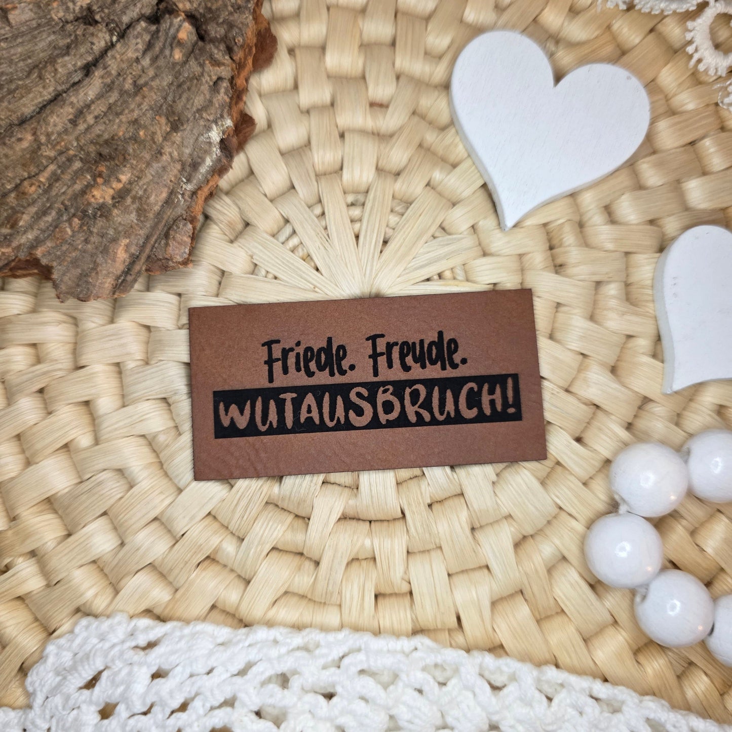 Kunstlederlabel „Friede. Freude. Wutausbruch!“ aus braunem Kunstleder (6 × 3 cm) mit schwarzer Gravur, dekorativ auf Bastuntergrund mit Holzrinde, weißen Dekoherzen und Häkelspitze arrangiert – humorvolles Label für selbstgenähte Kleidung, Kinderoutfits oder Accessoires mit Charakter.