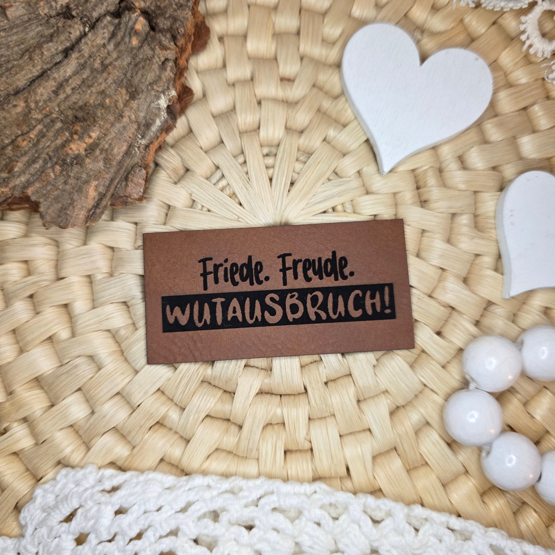 Kunstlederlabel „Friede. Freude. Wutausbruch!“ aus braunem Kunstleder (6 × 3 cm) mit schwarzer Gravur, dekorativ auf Bastuntergrund mit Holzrinde, weißen Dekoherzen und Häkelspitze arrangiert – humorvolles Label für selbstgenähte Kleidung, Kinderoutfits oder Accessoires mit Charakter.