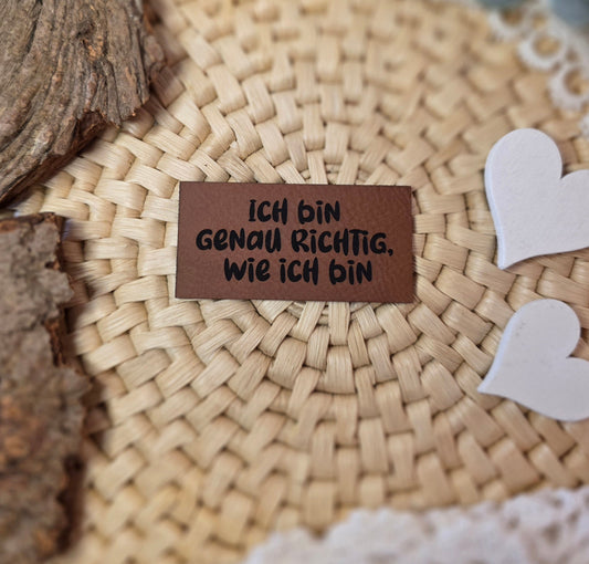 Kunstlederlabel „Ich bin genau richtig, wie ich bin“ aus braunem Kunstleder (6 × 3 cm) mit schwarzer Gravur auf Bastuntergrund, dekoriert mit Holzrinde, weißen Dekoherzen und Spitze – ein liebevolles Detail für selbstgenähte Kleidung oder Accessoires mit positiver Botschaft.