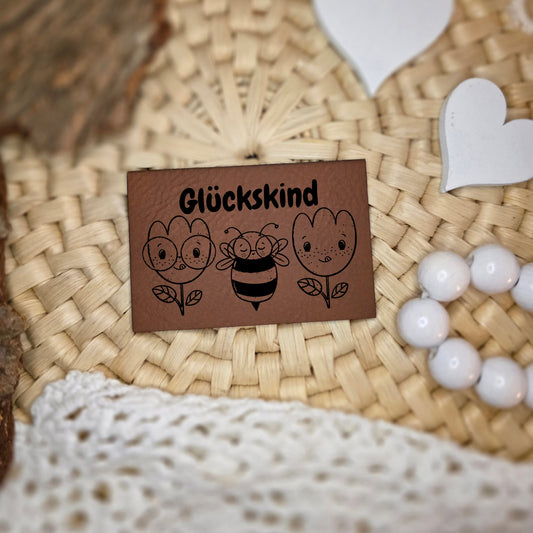 Kunstlederlabel „Glückskind“ 4×6 cm mit Schriftzug sowie zwei Blumen und Biene, schwarz graviert auf braunem Kunstleder, dekorativ auf Naturuntergrund fotografiert
