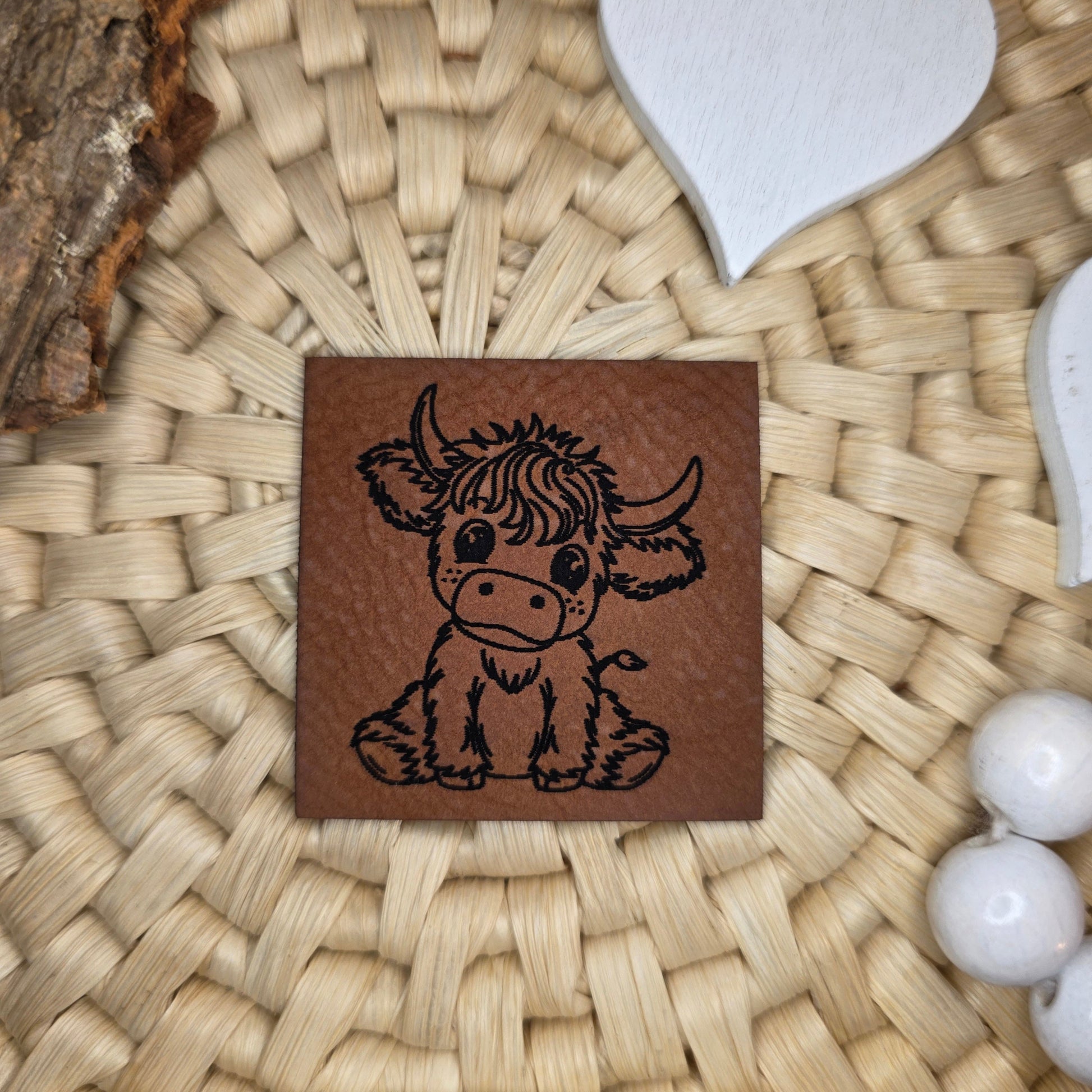 Quadratisches Kunstlederlabel „Hochlandrind“ aus braunem Kunstleder (4 × 4 cm) mit schwarzer Gravur – handgezeichnetes Katzengold-Design eines süßen Hochlandrinds, dekorativ auf Bastuntergrund mit Holzrinde, weißen Dekoherzen und Häkelspitze arrangiert – perfektes Label für selbstgenähte Kinderkleidung und Accessoires im Landhausstil.