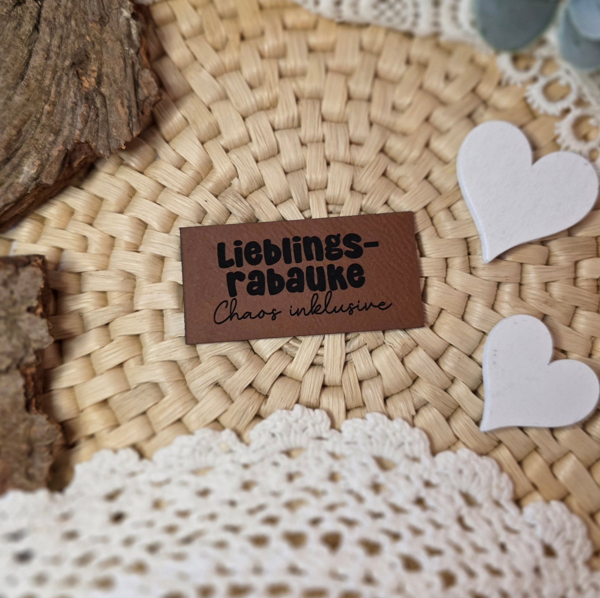 Kunstlederlabel „Lieblingsrabauke – Chaos inklusive“ aus braunem Kunstleder (6 × 3 cm) mit schwarzer Gravur, dekorativ platziert auf Bastuntergrund mit Holzrinde, weißen Dekoherzen und Häkelspitze – ein verspieltes Detail für selbstgenähte Kinderkleidung oder Accessoires.