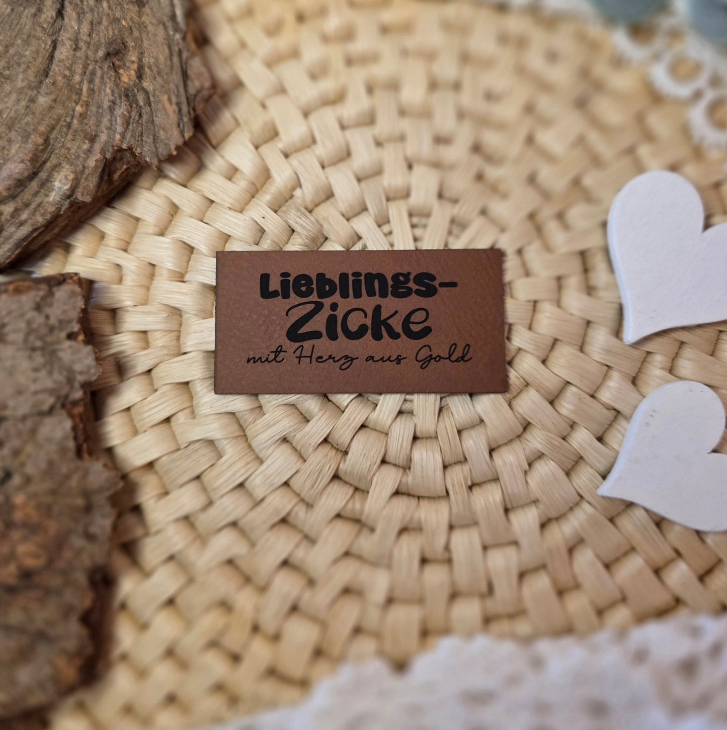 Kunstlederlabel „Lieblings-Zicke mit Herz aus Gold“ aus braunem Kunstleder (6 × 3 cm) mit schwarzer Gravur auf Bastuntergrund, umgeben von Holzrinde, weißen Dekoherzen und Spitze – ideal als witziges Detail für selbstgenähte Kleidung oder Accessoires.