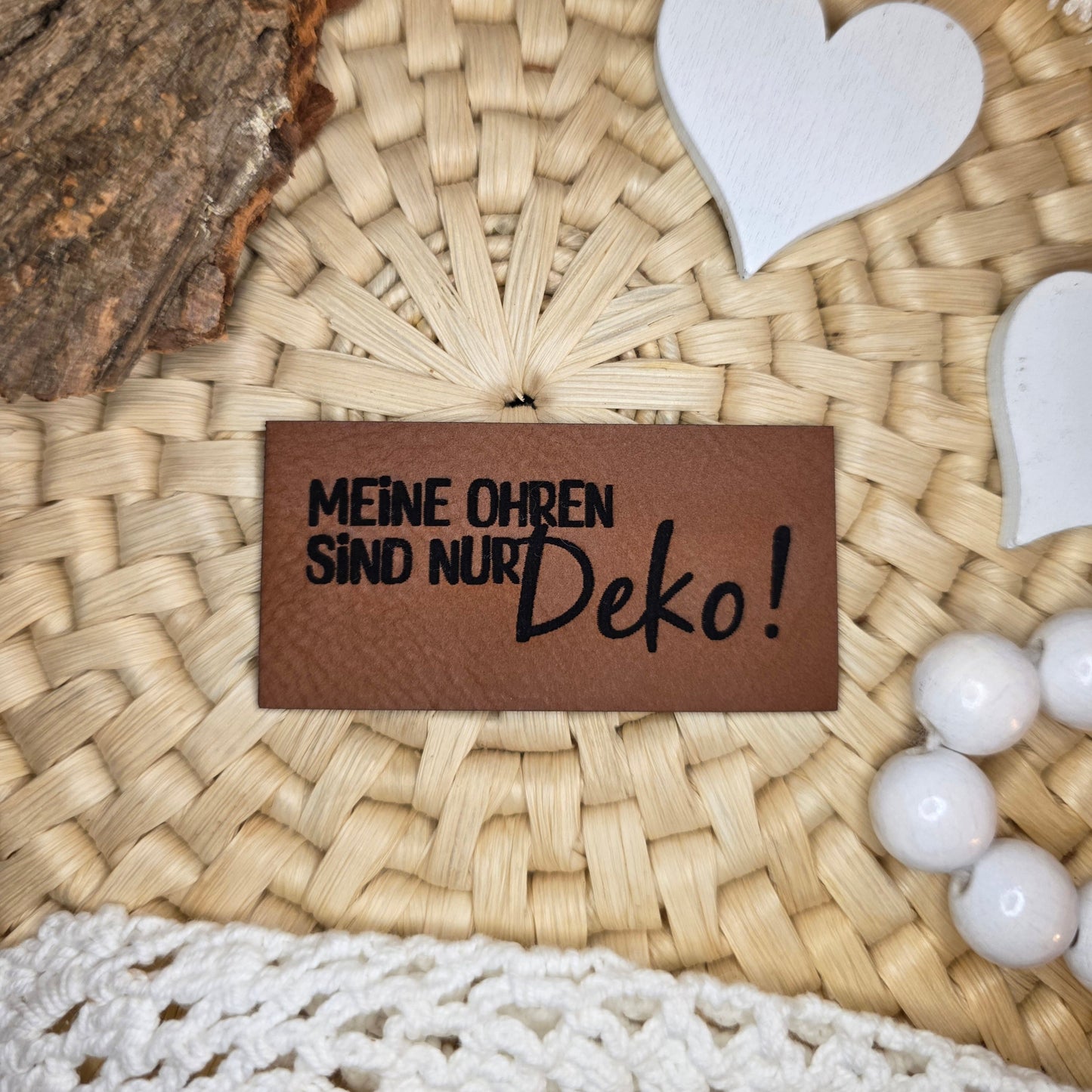 Kunstlederlabel „Meine Ohren sind nur Deko!“ aus braunem Kunstleder (6 × 3 cm) mit schwarzer Gravur, dekorativ auf Bastuntergrund mit Holzrinde, weißen Dekoherzen und Häkelspitze arrangiert – humorvolles Statement-Label für selbstgenähte Kleidung, Taschen oder Accessoires.