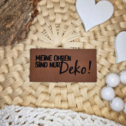 Kunstlederlabel „Meine Ohren sind nur Deko!“ aus braunem Kunstleder (6 × 3 cm) mit schwarzer Gravur, dekorativ auf Bastuntergrund mit Holzrinde, weißen Dekoherzen und Häkelspitze arrangiert – humorvolles Statement-Label für selbstgenähte Kleidung, Taschen oder Accessoires.