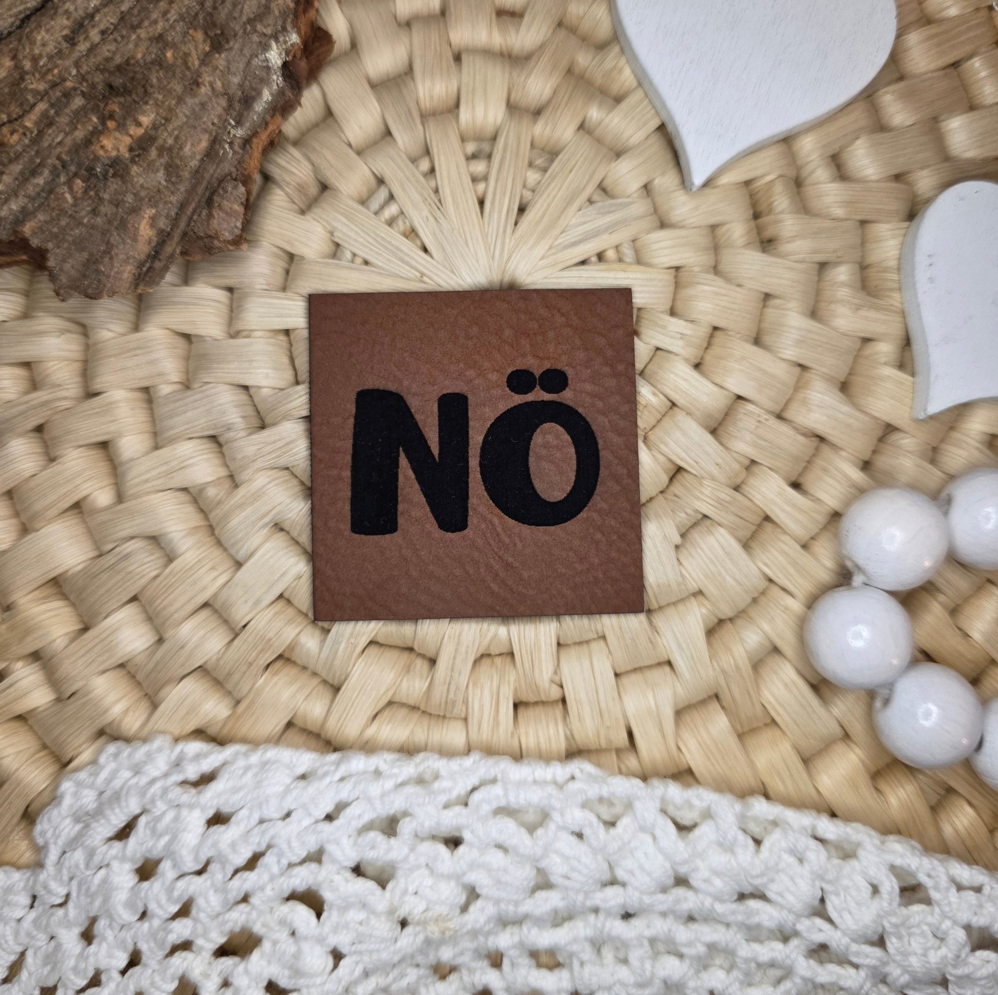 Quadratisches Kunstlederlabel „NÖ“ aus braunem Kunstleder (4 × 4 cm) mit schwarzer Gravur, dekorativ platziert auf Bastuntergrund mit Holzrinde, weißen Dekoherzen und Häkelspitze – minimalistisches Statement-Label für Kleidung, Accessoires oder Taschen mit Haltung.