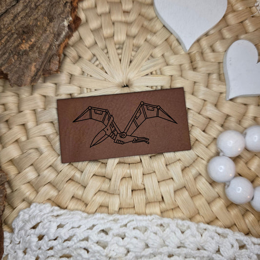 Rechteckiges Kunstlederlabel mit graviertem Robo-Flugsaurier im technischen Dino-Stil, Motivdesign von Katzengold-Designs, gefertigt von Stich & Schere