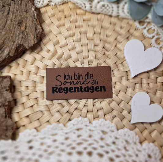 Kunstlederlabel „Ich bin die Sonne an Regentagen“ aus braunem Kunstleder (6 × 3 cm) mit schwarzer Gravur, dekoriert auf Bastuntergrund mit Holzrinde, weißen Dekoherzen und Spitze – ein detailverliebtes Label für Handmade-Kleidung und Accessoires mit positiver Botschaft.