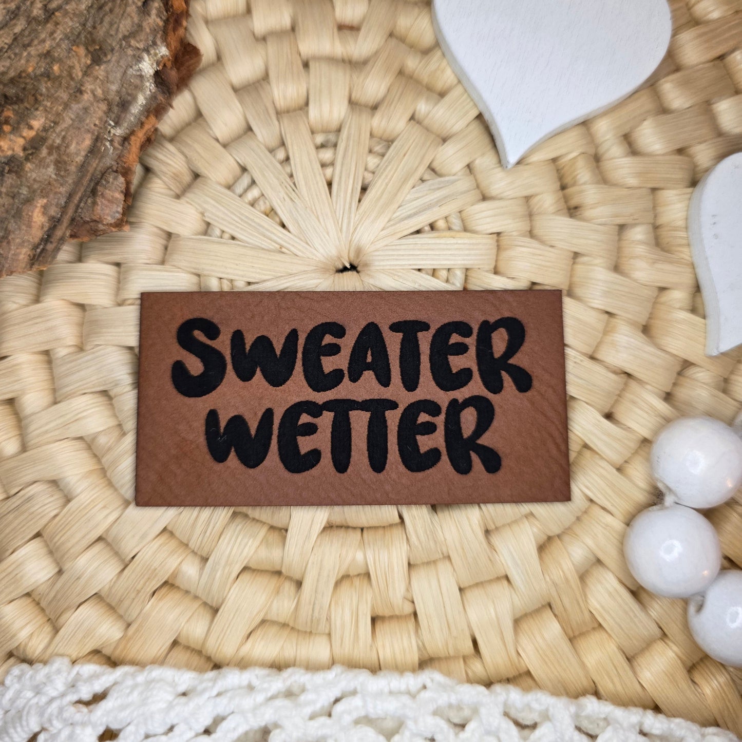 Kunstlederlabel „Sweater Wetter“ aus braunem Kunstleder (6 × 3 cm) mit schwarzer Gravur, dekorativ auf Bastuntergrund mit Holzrinde, weißen Dekoherzen und Häkelspitze arrangiert – stilvolles Label für selbstgenähte Hoodies, Pullis und gemütliche Herbstkleidung.