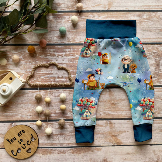 Handgemachte Baby-Pumphose „Fliegende Träume“ aus Bio-French Terry mit verträumtem Ballon-Design, kombiniert mit dunkelblauen Bündchen, dekorativ auf Holzuntergrund arrangiert.