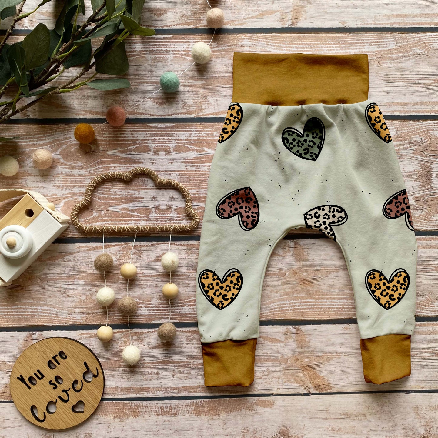 Handgemachte Baby-Pumphose „Leo Herzen“ aus Bio-French Terry mit verspielten Leoparden-Herzen auf hellem Grund und senfgelben Bündchen, dekorativ auf Holzuntergrund arrangiert.