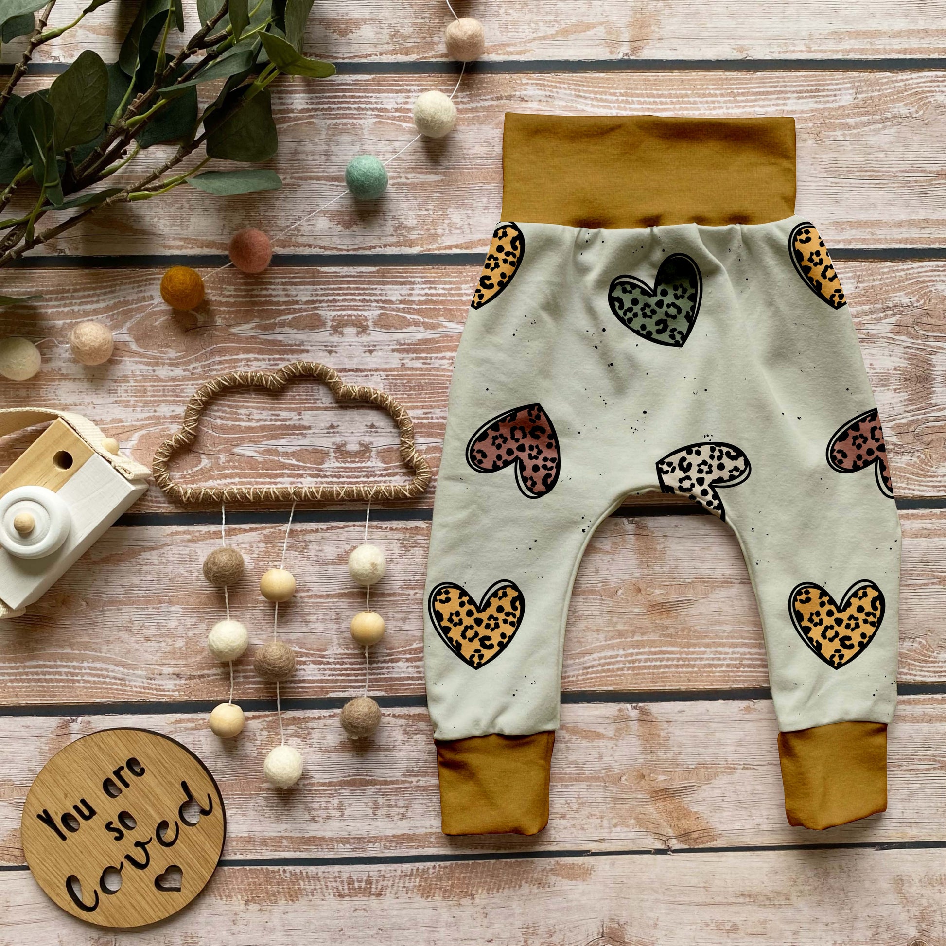 Handgemachte Baby-Pumphose „Leo Herzen“ aus Bio-French Terry mit verspielten Leoparden-Herzen auf hellem Grund und senfgelben Bündchen, dekorativ auf Holzuntergrund arrangiert.
