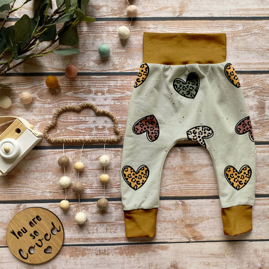 Handgemachte Baby-Pumphose „Leo Herzen“ aus Bio-French Terry mit verspielten Leoparden-Herzen auf hellem Grund und senfgelben Bündchen, dekorativ auf Holzuntergrund arrangiert.
