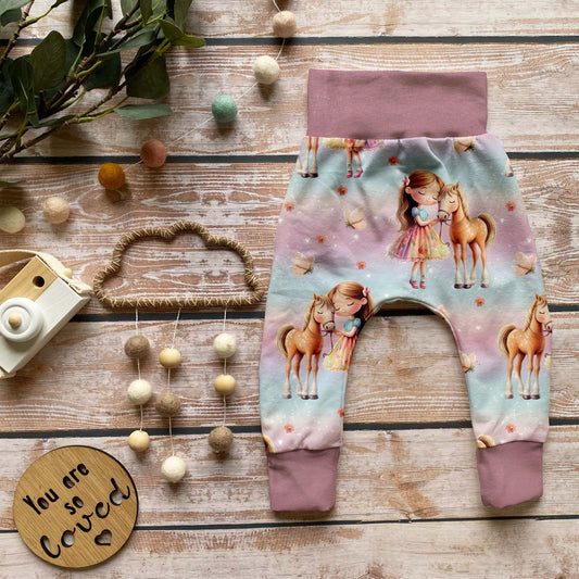 Handgemachte Baby-Pumphose „Mädchen mit Pferd“ mit Bündchen in Altrosa aus Bio-French Terry mit süßem Pferde-Design und weißen Bündchen, auf Holzuntergrund dekorativ arrangiert.