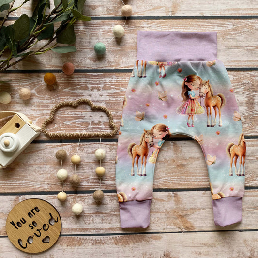 Handgemachte Baby-Pumphose „Mädchen mit Pferd“ mit Bündchen in Lavendel aus Bio-French Terry mit süßem Pferde-Design und weißen Bündchen, auf Holzuntergrund dekorativ arrangiert.
