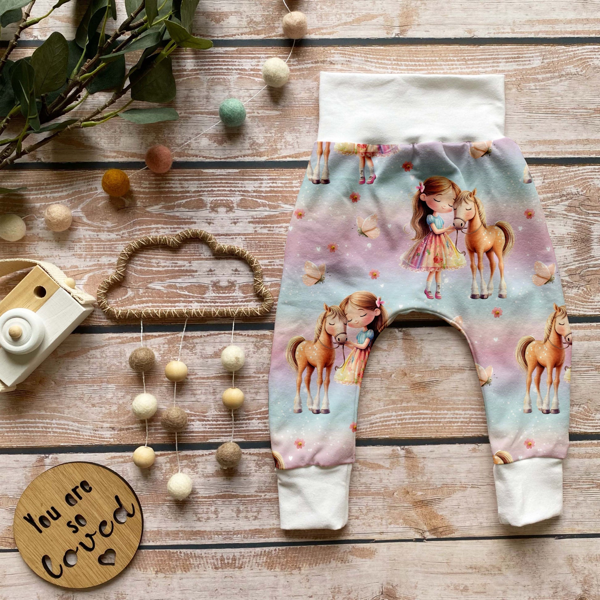 Handgemachte Baby-Pumphose „Pferdemädchen“ aus Bio-French Terry mit süßem Pferde-Design und weißen Bündchen, auf Holzuntergrund dekorativ arrangiert.