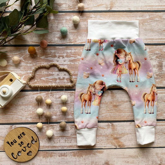 Handgemachte Baby-Pumphose „Pferdemädchen“ aus Bio-French Terry mit süßem Pferde-Design und weißen Bündchen, auf Holzuntergrund dekorativ arrangiert.