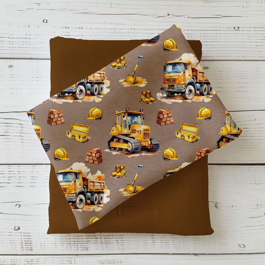 Mockup: Stoff „Baustellen-Abenteuer“ – gelbe Baufahrzeuge (Bagger, Bulldozer, Kipplaster) mit Helmen, Stiefeln, Ziegeln & Werkzeug auf taupefarbenem Hintergrund; Stoffkachel diagonal auf Uni in Cognac/Braun als Kombistoff.