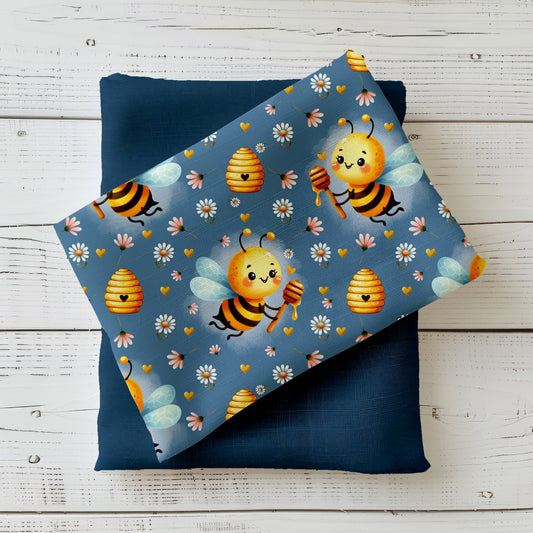 Mockup: Stoff „Bee Happy“ – niedliche Bienen mit Honiglöffel, Bienenkörbe, Gänseblümchen & goldene Herzen auf taubenblauem Hintergrund; Stoffkachel diagonal auf passendem Uni als Kombistoff.