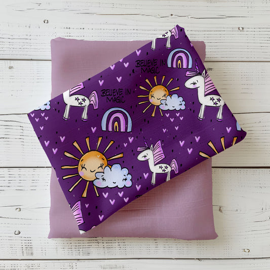 Mockup: Stoff „Believe in Magic – Einhorn“ in Lila mit weißen Einhörnern (rosa/lila Mähne), Regenbögen, Sonne und Wolke, vielen kleinen Herzchen und dem Schriftzug „Believe in Magic“; Stoffstück liegt diagonal auf fliederfarbenem Kombistoff, fotografiert auf weißer Holzfläche.