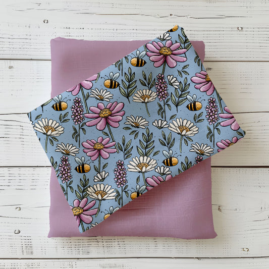 Gefalteter „Bunte Wiese“ Stoff von Katzengold-Designs – farbenfrohe Blumen, Blätter und kleine Bienen auf blaugrünem Hintergrund, dekorativ auf fliederfarbenem Kombistoff präsentiert