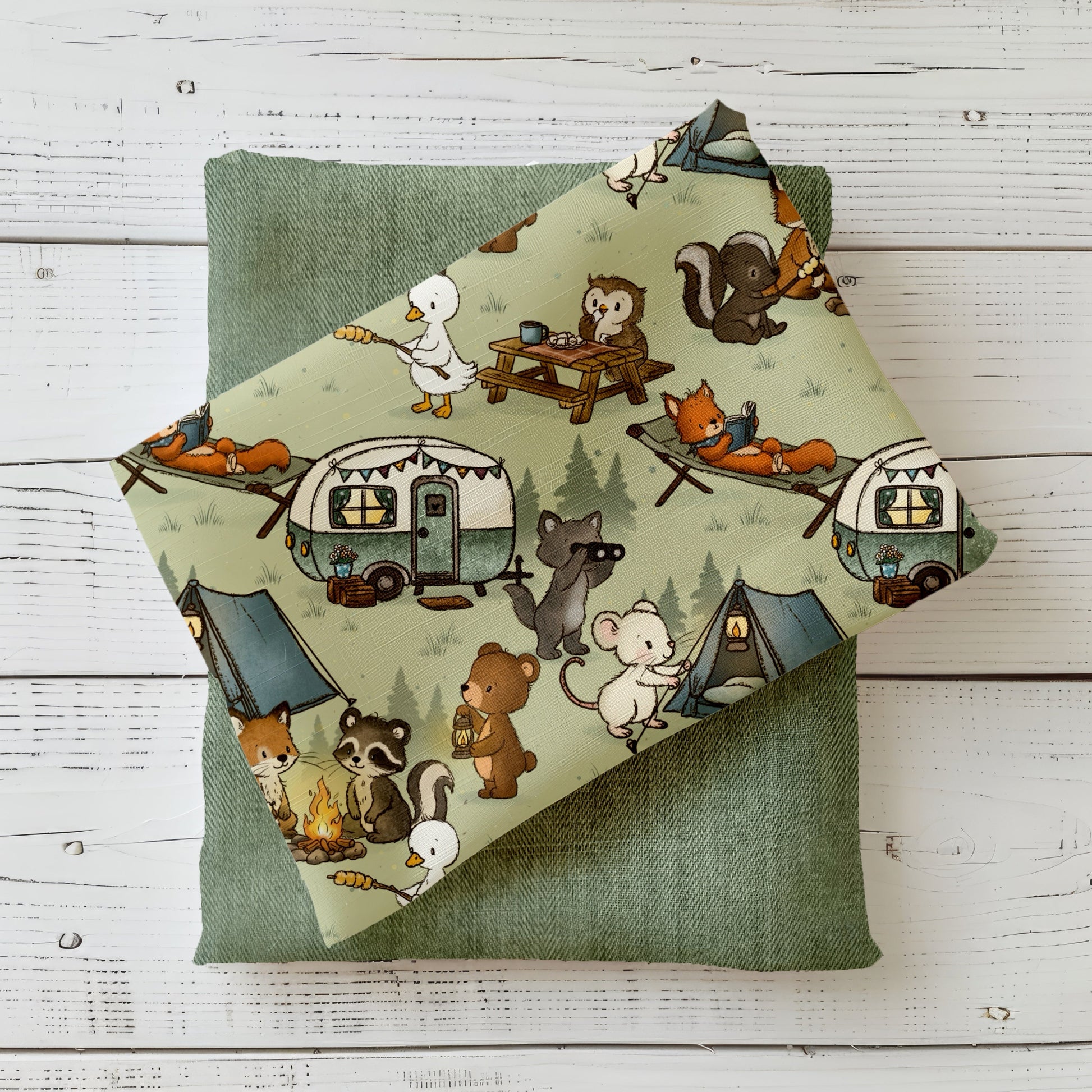 Dekorativ fotografierter Stoff mit dem Design „Campingfreunde“, auf einem Kissen drapiert. Der Stoff zeigt die liebevoll gezeichneten Camping-Szenen mit Waldtieren wie Fuchs, Waschbär und Bär. Sichtbar sind Details wie ein Lagerfeuer, Zelte und ein kleiner Wohnwagen. Der Stoff liegt auf einem grün strukturierten Kissen auf hellem Holzuntergrund, wodurch die natürlichen Farben besonders gut zur Geltung kommen. Die Szene wirkt warm, gemütlich und lädt zum Wohlfühlen ein.