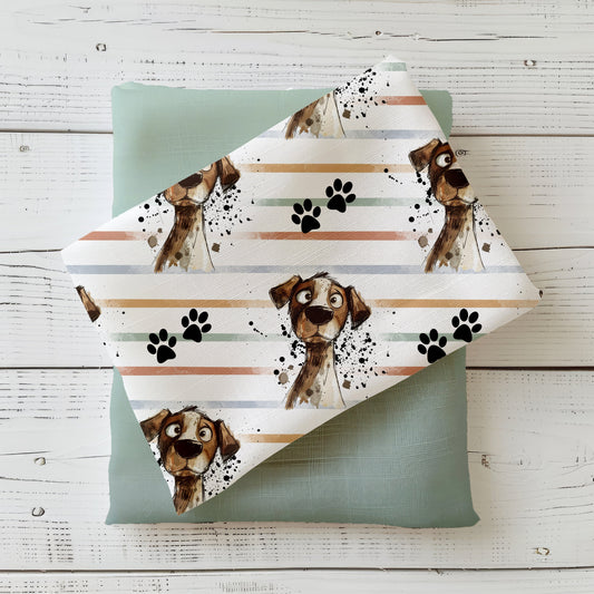 Mockup: Stoff „Crazy Dog“ – gezeichneter Hundekopf mit witzigem Gesichtsausdruck auf Streifen in Salbei, Sand, Pfirsich und Grau; schwarze Pfotenabdrücke; Stoffstück diagonal auf salbeigrünem Kombistoff, fotografiert auf weißer Holzfläche.