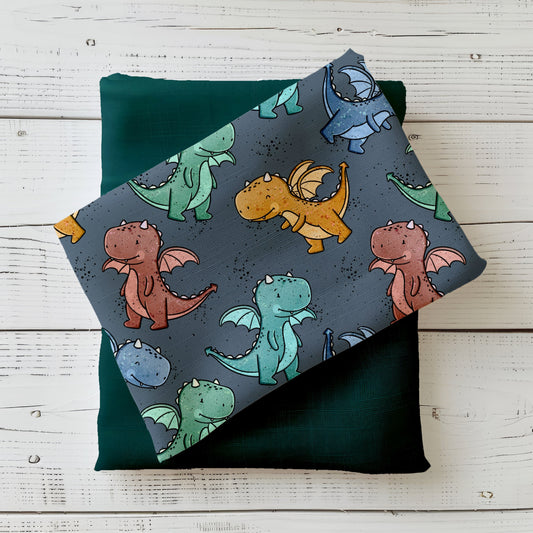 Gefalteter Stoff mit niedlichen Drachenmotiven in Grün-, Blau- und Rosttönen auf dunklem Hintergrund, dekorativ auf Holzuntergrund arrangiert.