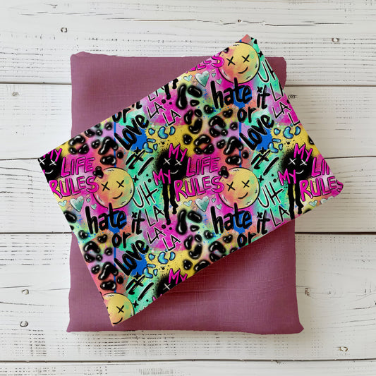 Mockup: Stoff „Hate it or Love it“ – Graffiti-Schriftzüge, Smileys, Drips & Leo-Sprenkel in Neon-Pink, Lila, Türkis, Gelb & Lime auf dunklen Akzenten; Stoffkachel diagonal auf passendem Uni in Mauve/Lila als Kombi.