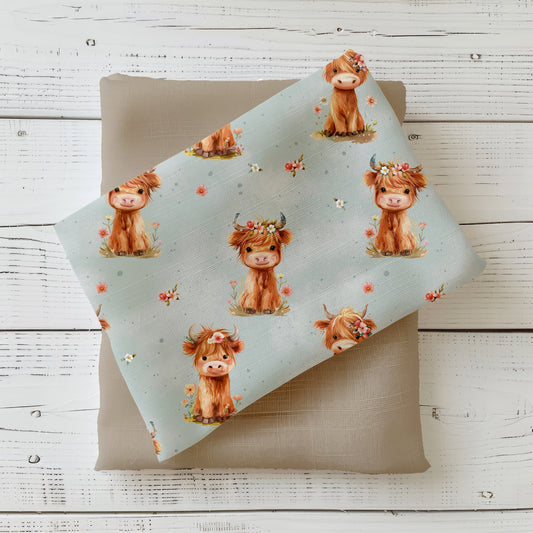 Mockup: Stoff „Highland Cow“ mit niedlichen Hochlandrindern, teils mit Blumenkranz, auf hellem Mint mit Punkten; Stoffstück liegt diagonal auf sandfarbenem Kombistoff, fotografiert auf weißer Holzfläche.