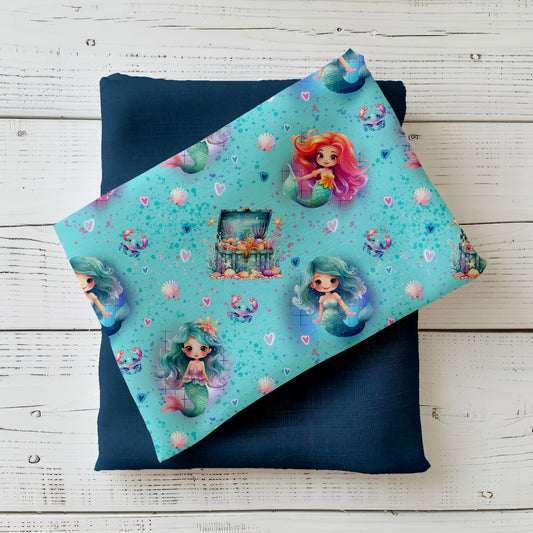 Mockup: Stoff „Kleine Meerjungfrau“ – niedliche Meerjungfrauen in Türkis & Koralle mit Herzen, Muscheln, Krebsen und Schatztruhe auf aquablauem Hintergrund; Stoffkachel diagonal auf passendem Uni als Kombi.