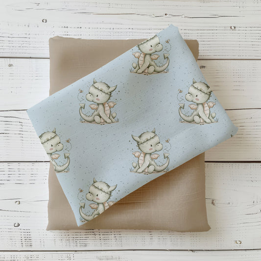 Mockup: Stoff „Kleiner Drache“ – kleiner Babydrache mit Flügelchen und Biene auf hellblauem Hintergrund; Stoffstück diagonal auf sandbeigem Kombistoff, fotografiert auf weißer Holzfläche.