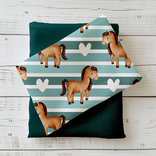 Gefalteter Stoff „Ponyliebe“ mit niedlichen Ponys auf gestreiftem Hintergrund, drapiert auf einfarbigem Kombistoff – Produktmockup auf hellem Holzhintergrund.